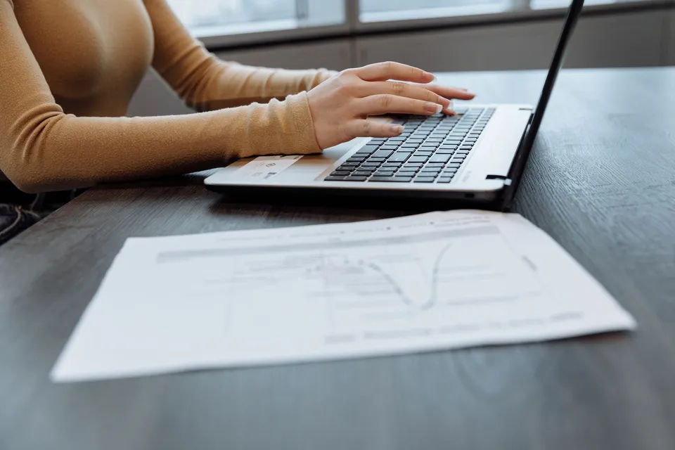 Close-up of a person's hands typing on a laptop at a desk with documents. Ideal for office, work, and technology themes.