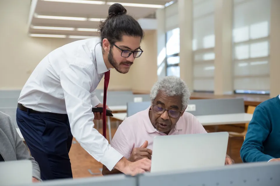 A young man helps a senior adult with laptop use, emphasizing tech education.